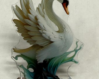 May include: A clear acrylic pin depicting a white swan with its wings spread, a curved neck, and an orange beak. The swan is set against teal and green water. The pin has two silver clasps on the back.