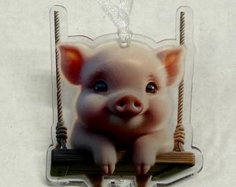 May include: A clear acrylic ornament depicting a smiling piglet on a swing. The piglet is pink with dark eyes and is hanging onto a wooden swing. The swing is suspended by rope and has a white ribbon for hanging.