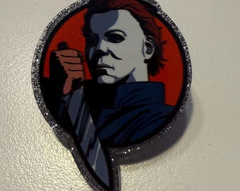 May include: A pin badge depicting Michael Myers from the Halloween film franchise. The image shows Myers holding a knife against a red backdrop, framed by a silver glitter border.