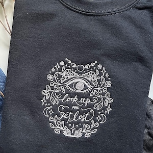 Könnte beinhalten: Schwarzes Sweatshirt mit weißem Stickdesign. Das Design zeigt ein Auge, florale Elemente und die Worte "look up and get lost". Das Sweatshirt ist gefaltet, mit schwarzen Schuhen im Hintergrund.
