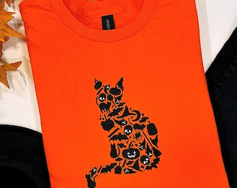 May include: Bright orange t-shirt featuring a black cat silhouette composed of Halloween-themed icons. The cat design includes bats, pumpkins, skulls, and other spooky elements. The shirt is a crew neck style.
