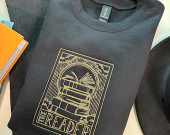 Puede incluir: Sudadera negra con un diseño bordado en dorado que presenta una pila de libros y el texto "THE READER". La sudadera está doblada sobre unos vaqueros oscuros. Un sombrero negro y flores naranjas están en el fondo.