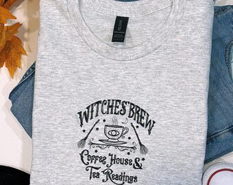 Puede incluir: Una camiseta gris jaspeada con el texto "Witches Brew Coffee House & Tea Readings" en escritura negra. El diseño incluye una taza de café con un ojo, dos escobas y adornos decorativos. La camiseta está doblada sobre unos vaqueros azules y zapatillas blancas.
