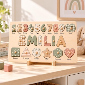 May include: A rectangular wooden educational puzzle board featuring the name EMILIA in large letters, numbers one through ten, and six geometric shapes including a square, triangle, circle, star, hexagon, and heart, all with small wooden knobs.
