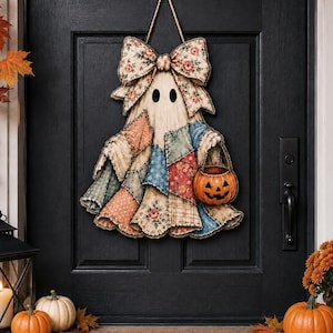May include: A hanging ghost decoration featuring a large floral patterned bow at the top and a patchwork-style body made of various fabric prints in shades of blue, red, and beige. The ghost holds a small orange jack-o'-lantern bucket in one hand.