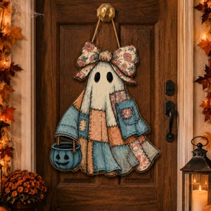 May include: A decorative wall hanging shaped like a ghost, featuring a patchwork design in shades of blue, tan, and floral patterns. The ghost wears a large matching floral bow on its head and holds a blue jack-o'-lantern bucket by a rope handle.