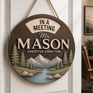 Op de afbeelding: Een rond houten deurbordje dat aan een touw hangt, met een donkerbruine achtergrond en een landschap van bergen, dennenbomen en een rivier. De tekst luidt IN A MEETING, Mr. MASON en CREATIVE DIRECTOR in gelaagde, lichtgekleurde letters.