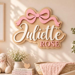 Puede incluir: Un letrero de pared de madera con el nombre Juliette en letras cursivas blancas grandes y el nombre ROSE en letras de bloque rosas. Un lazo rosa está sujeto a la parte superior de la placa de madera clara, que tiene un borde festoneado.