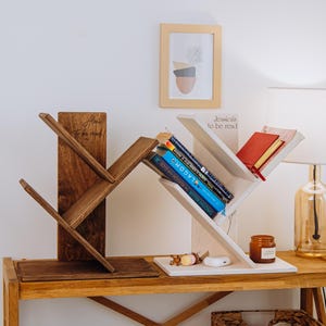 Puede incluir: Dos estanterías de madera de diferentes estilos se encuentran sobre una mesa de madera. Una es de color marrón oscuro y con forma de árbol, la otra es blanca e inclinada. Los libros se exhiben en las estanterías y una imagen enmarcada cuelga de la pared.