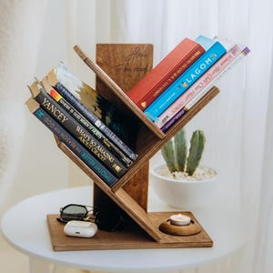May include: A wooden bookshelf in a tree-like design, holding several books with colorful spines. The bookshelf is brown and sits on a white table. Sunglasses, a wireless earbud case, and a small candle are also on the table. The text "Alexis To be read" is on the bookshelf.
