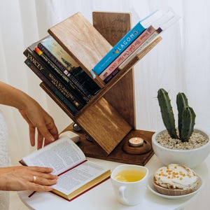 Puede incluir: Una estantería de madera con estantes angulados que sostienen varios libros. Una persona sostiene un libro abierto con una cubierta roja. Una mesa blanca tiene una taza de té, una magdalena y un pequeño cactus en una maceta blanca. Una vela está encendida.