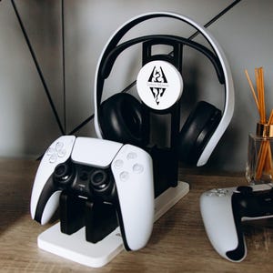 以下が含まれることがあります： 白と黒のゲーミングヘッドセットと2つの白と黒のゲームコントローラーが木製の表面に表示されています。ヘッドセットは、「James Mason」と書かれた白い円形のロゴが付いた黒いスタンドにあります。コントローラーは白い充電スタンドにあります。