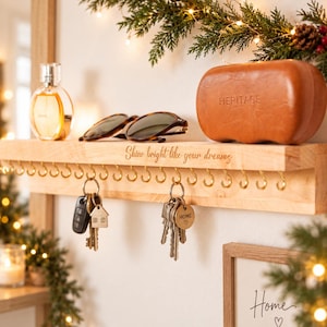 Op de afbeelding: Een lichtgekleurde houten wandplank met een rij goudkleurige metalen haken. Op de voorkant van de plank staat de tekst 'Shine bright like your dreams' gegraveerd. Aan de haken hangen twee sleutelbossen, één met een huisvormige bedel en de andere met een rond label met de tekst 'HOME'.