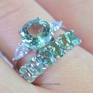 May include: Two rings with gemstones. The engagement ring features a large, round, light green gemstone with two pear-shaped white gemstones on either side. The wedding band has multiple oval-shaped light green gemstones. The rings are set in silver.