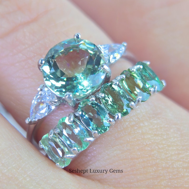 May include: Two rings with gemstones. The engagement ring features a large, round, light green gemstone with two pear-shaped white gemstones on either side. The wedding band has multiple oval-shaped light green gemstones. The rings are set in silver.