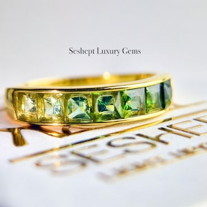 May include: A gold band ring with a row of green and clear square-shaped gemstones. The ring is set on a white surface with the text "Seshept Luxury Gems" printed on it.