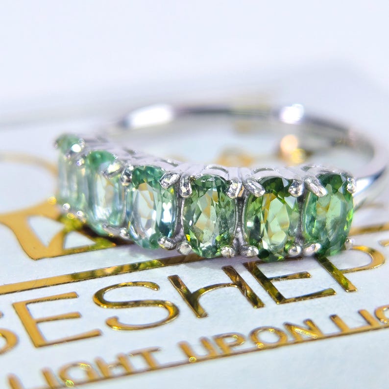 May include: A silver ring featuring a row of oval-cut, light green gemstones. The ring is set against a white background with gold lettering that reads "LIGHT UPON LIGHT". The gemstones are evenly spaced and reflect light.