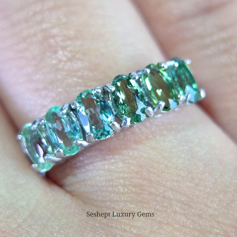 May include: A close-up of a silver ring featuring a row of oval-cut, teal-green gemstones. The ring is set against a blurred background, with the text "Seshept Luxury Gems" visible at the bottom. The gemstones have a vibrant, sparkling appearance.