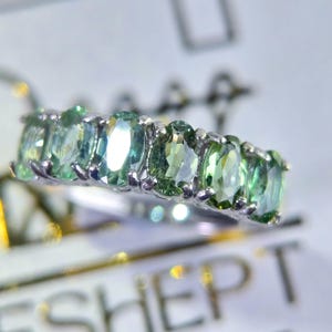 May include: A silver ring featuring a row of oval-cut, light green gemstones. The gemstones are set closely together, creating a continuous band of color. The ring is displayed on a white surface, highlighting the sparkle of the stones.