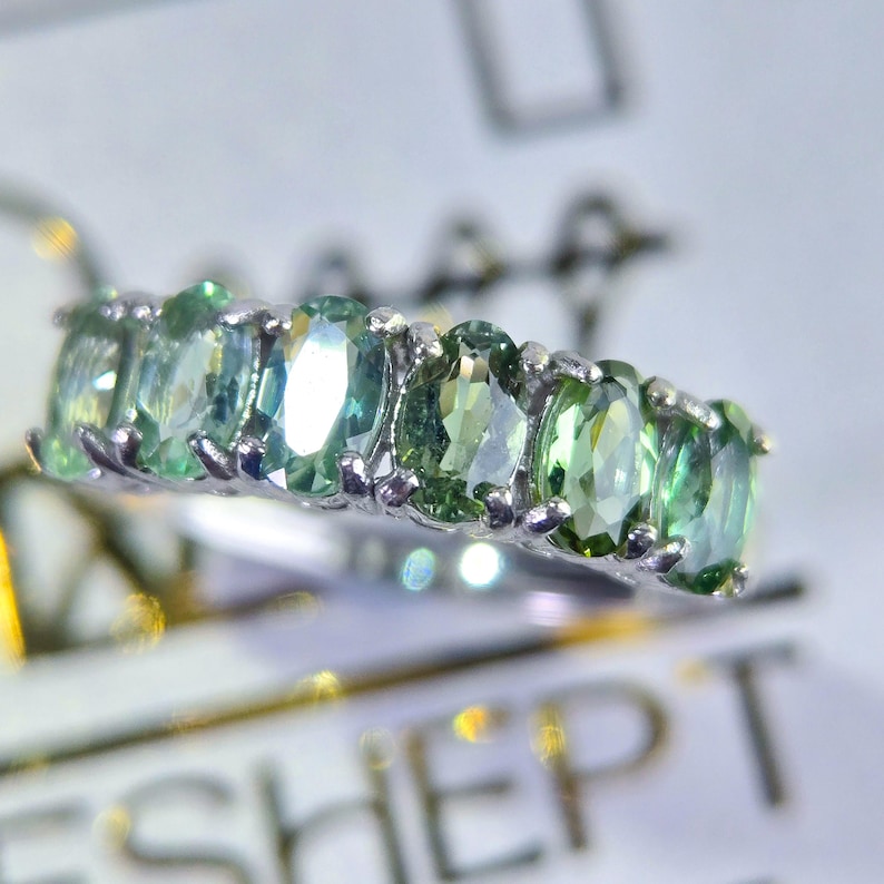 May include: A silver ring featuring a row of oval-cut, light green gemstones. The gemstones are set closely together, creating a continuous band of color. The ring is displayed on a white surface, highlighting the sparkle of the stones.