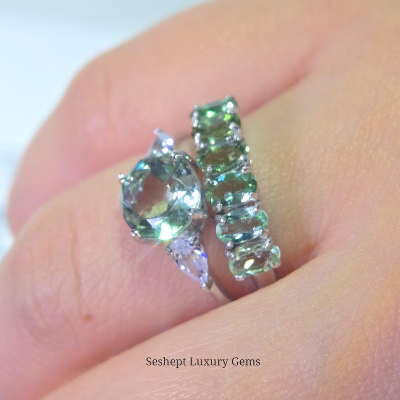 May include: Two rings with green gemstones. One ring features a large, round-cut green gemstone with smaller, clear accent stones. The second ring has a band of oval-cut green gemstones. The rings are set in silver. Text: Seshept Luxury Gems.