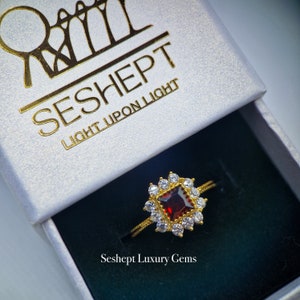 May include: A gold ring featuring a red gemstone encircled by smaller white gemstones. The ring is presented in a white box with the inscription "Seshept Light Upon Light" and "Seshept Luxury Gems".