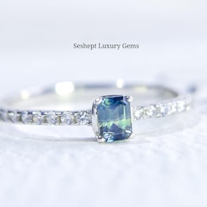 Op de afbeelding: Een zilveren ring met een rechthoekige, veelkleurige edelsteen in het midden. De band is versierd met kleine, heldere stenen. De tekst "Seshept Luxury Gems" is zichtbaar boven de ring.