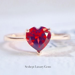 Puede incluir: Un anillo de oro con una piedra preciosa roja en forma de corazón. El anillo está sobre un fondo blanco.