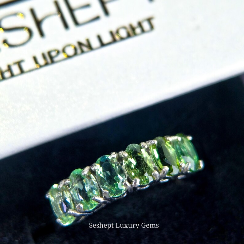 May include: A silver ring featuring a row of oval-cut, light green gemstones. The ring is set against a black background, with the text "Seshept Luxury Gems" visible. The ring is a piece of jewelry.