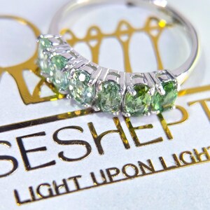 May include: A silver ring with five oval-cut, light green gemstones set in a row. The ring is on a white surface with gold text that reads "SESH" and "LIGHT UPON LIGHT". The gemstones have a slight gradient in color.