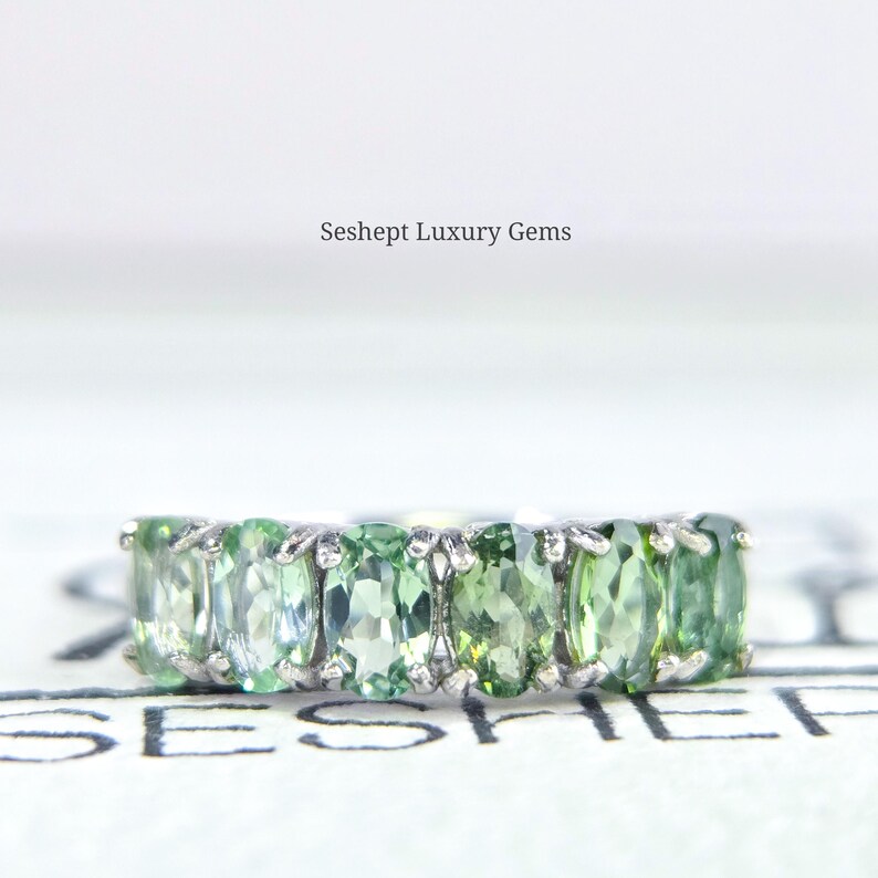 May include: A silver ring featuring a row of oval-cut, light green gemstones. The ring is set against a white background, with the text "Seshept Luxury Gems" visible. The gemstones have a vibrant, verdant hue.