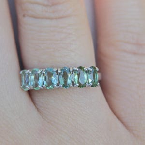May include: A silver ring featuring a row of oval-cut gemstones in shades of blue and green. The gemstones are set closely together, creating a continuous band of color. The ring is shown on a finger.
