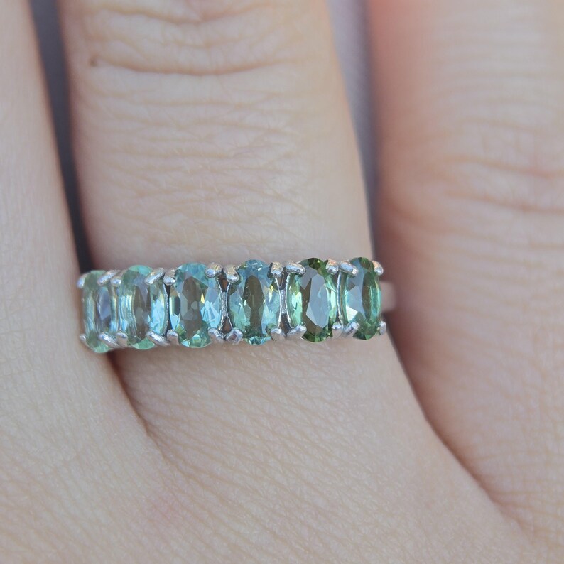 May include: A silver ring featuring a row of oval-cut gemstones in shades of blue and green. The gemstones are set closely together, creating a continuous band of color. The ring is shown on a finger.
