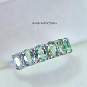 May include: A silver ring featuring a row of oval-cut, light green gemstones. The gemstones are set closely together, creating a continuous band of color. The text "Seshept Luxury Gems" is visible.