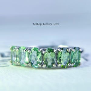 May include: A silver ring featuring a row of oval-cut, light green gemstones. The gemstones are set closely together, creating a continuous band of color. The ring is set against a soft, blurred background. The text "Seshept Luxury Gems" is visible.