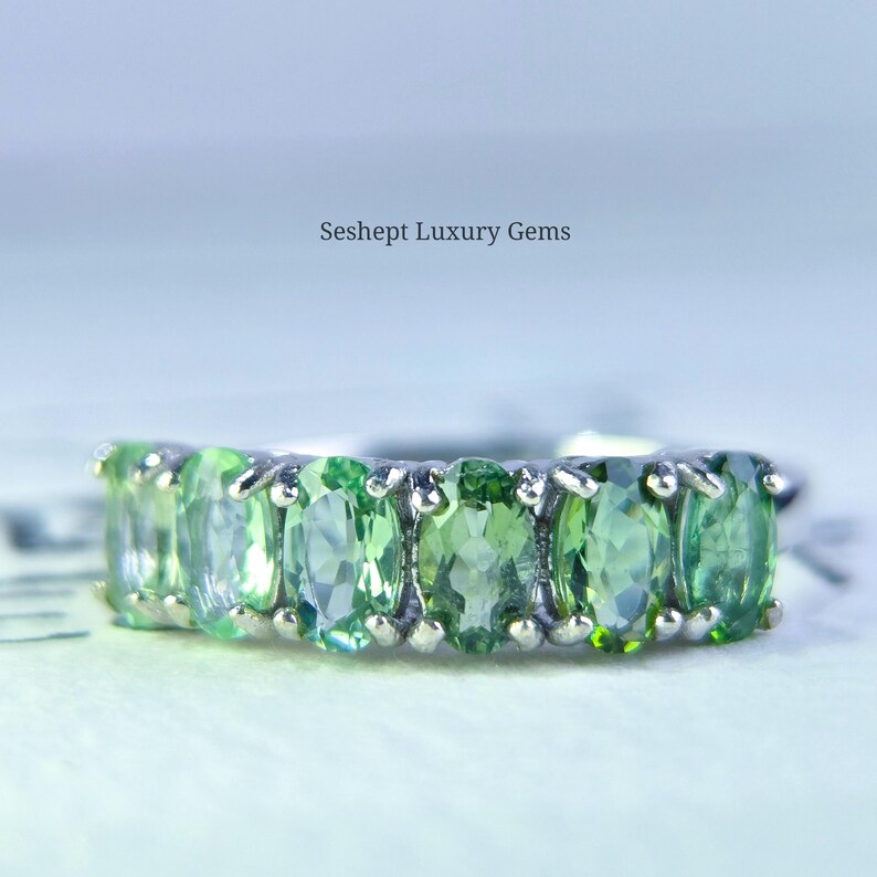 May include: A silver ring featuring a row of oval-cut, light green gemstones. The gemstones are set closely together, creating a continuous band of color. The ring is set against a soft, blurred background. The text "Seshept Luxury Gems" is visible.