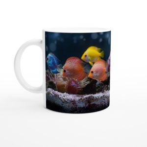 Könnte beinhalten: Weiße Keramiktasse mit einem bunten Bild von Fischen, die in einem Aquarium schwimmen. Die Fische sind orange, gelb und blau mit schwarzen Markierungen. Der Hintergrund ist dunkelblau mit Blasen.