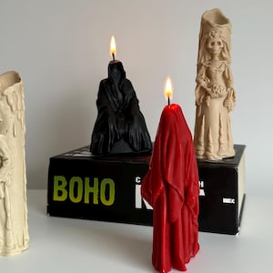 Puede incluir: Un conjunto de velas encendidas con formas únicas. Una vela es un esqueleto con traje, otra es una figura con capa negra, una figura roja y una novia esqueleto. Las velas están sobre una superficie blanca.