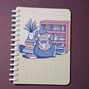 May include: A spiral-bound notebook featuring a cartoon mouse reading. The mouse is sitting on a blue beanbag, surrounded by books and a bookshelf. A potted plant is also shown. The notebook has a grid pattern.