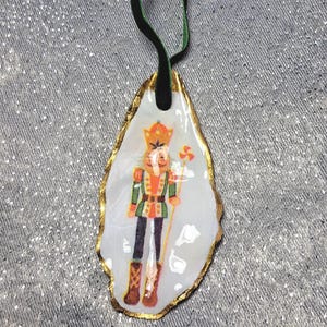 May include: A Christmas ornament with a nutcracker design. The ornament is made from a white, irregularly shaped material with a gold-coloured trim. The nutcracker is depicted in a colourful uniform, holding a staff. A black and green ribbon is attached for hanging.