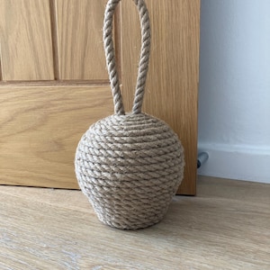 May include: A beige rope doorstop with a rounded base and a looped handle. The doorstop is made of tightly wound rope, creating a textured surface. It is placed on a light-coloured wooden floor.