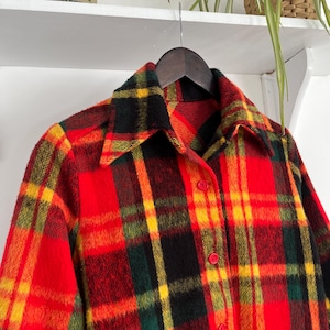 May include: A vintage plaid shirt in red, yellow, green, and black. The shirt is button-down with a classic collar, hanging on a wooden hanger. The fabric has a textured appearance. The shirt's pattern is a bold, colourful check.
