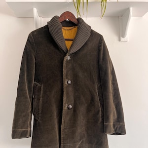May include: A dark olive green corduroy coat featuring a wide ribbed shawl collar, a mustard yellow interior lining, and three large dark buttons down the front. The coat has long sleeves and two side pockets, hanging on a wooden hanger.