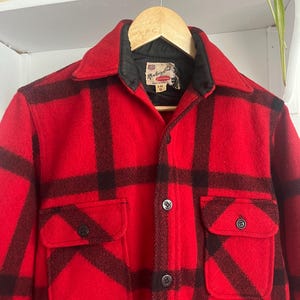 May include: A red and black plaid wool jacket with two front pockets and black buttons. The jacket is hanging on a wooden hanger. The label reads "Monterey Club".