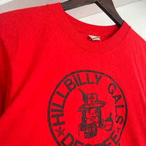 May include: A bright red short-sleeved t-shirt featuring a black circular graphic on the chest. The graphic contains the text "HILLBILLY GALS DEGREE" surrounding an illustration of a person wearing a hat and holding a bottle.