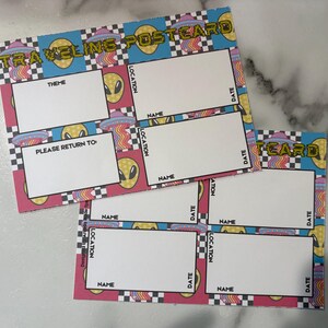 May include: Two colorful postcards with an alien theme. The postcards have a pink, blue, and yellow color scheme with the words "TRAVELING POSTCARD" and "CARD" printed on them. Each postcard has spaces for "THEME", "NAME", "LOCATION", and "DATE".