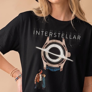 May include: A black short-sleeve t-shirt featuring the word INTERSTELLAR in white text above a graphic of a wristwatch with a black hole dial. Below the watch, a small illustration shows two figures looking upward at the watch face.