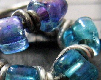 Puede incluir: Un brazalete envuelto en alambre plateado con cuentas de vidrio azul y morado iridiscentes. Las cuentas son redondas y tienen un acabado liso y pulido.
