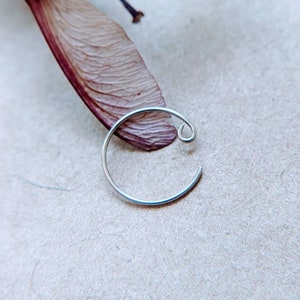 Puede incluir: Un aro de plata para la oreja con un pequeño círculo abierto en la parte superior. El aro está sobre una superficie de color marrón claro.