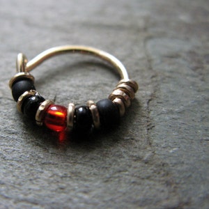 Puede incluir: Un aro de nariz de oro con cuentas negras y rojas. El aro está hecho de alambre fino y tiene un diseño pequeño y delicado.